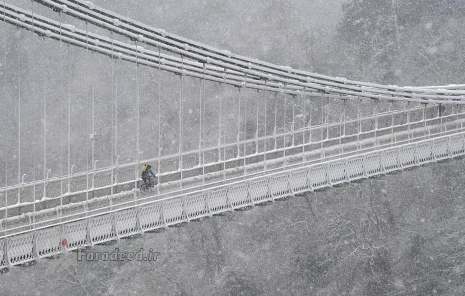 یک دوچرخه سوار از روی پل معلق کلیفتون و آون گورگ در جنوب غربی بریتانیا عبور می کند. برف به شدت در حال باریدن است.